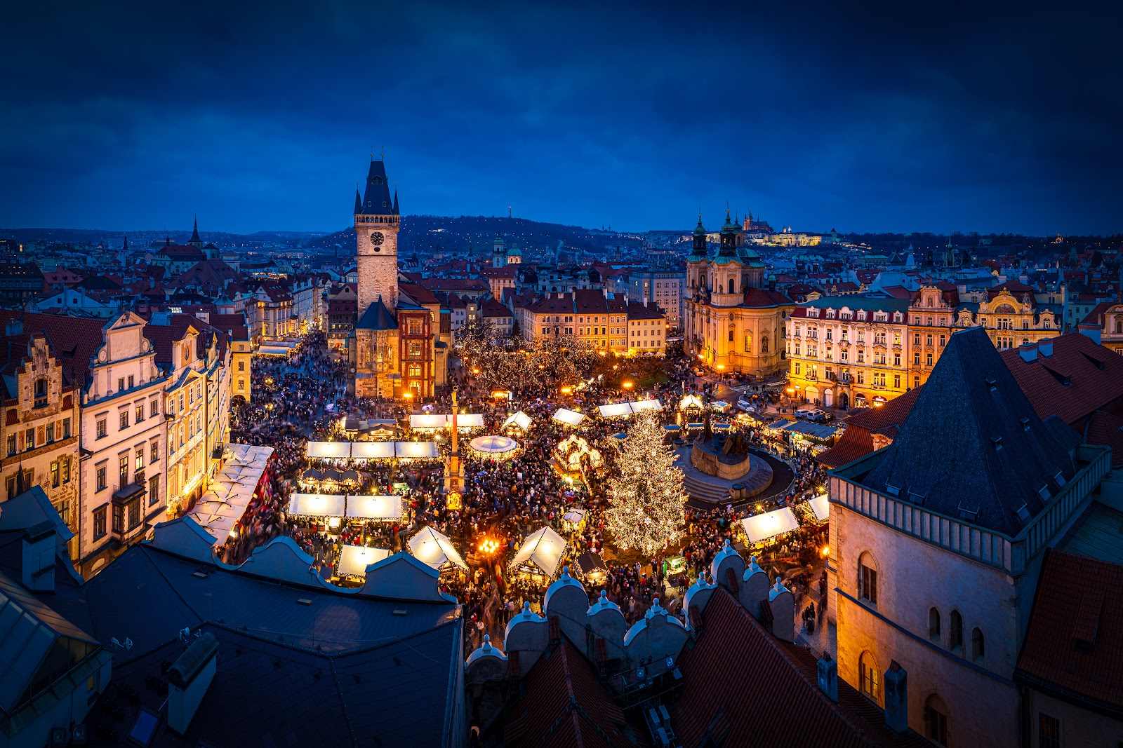 Old Town Square seen from above in the evening, market stalls lit between the roofs of the Old Town.