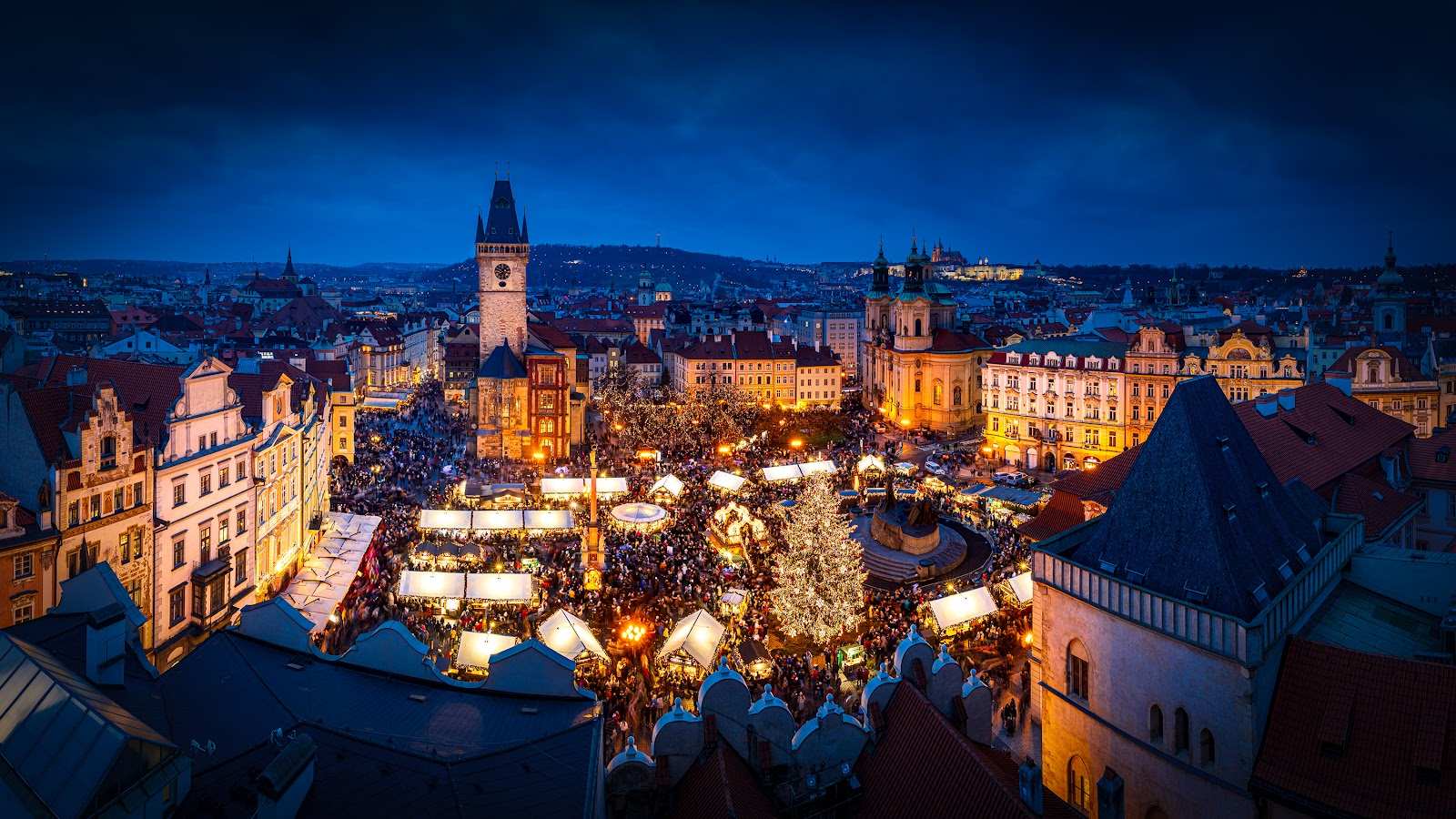 The Christmas tree and market on Old Town Square at dusk, with the Old Town Hall tower and Prague Castle behind.